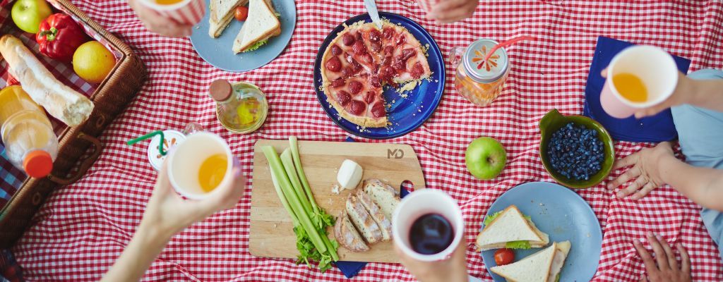 ChatGPT zei: Picknicksetting op een rood-wit geruit kleed met sandwiches, fruit, taart en drankjes, omringd door uitgestoken handen die bekers omhoog houden.