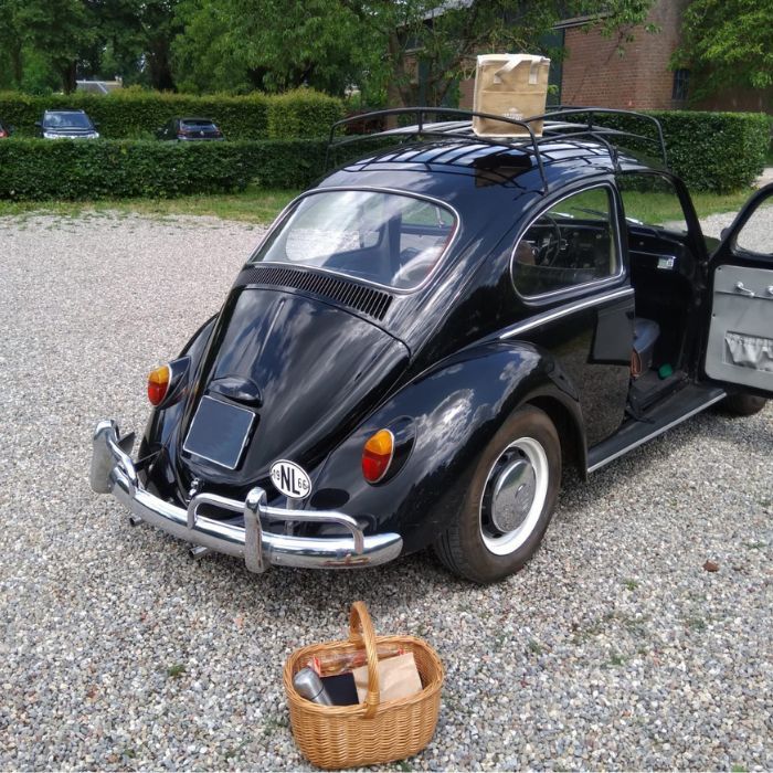 Zwarte klassieke Volkswagen Kever op een grindparkeerplaats met een kartonnen doos op het imperiaal en een open portier; op de grond staat een rieten picknickmand met inhoud.