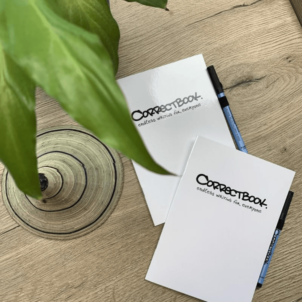 Twee witte Correctbook-schriften met zwarte opschrift en blauwe markers op een houten tafel, gedeeltelijk zichtbaar naast een plant en een decoratieve schaal.