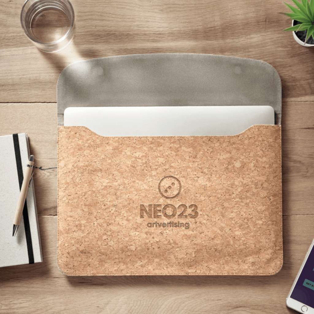 Kurken gepersonaliseerde laptophoes met zichtbaar ingestoken laptop, liggend op een houten bureau naast een notitieboek, een pen, een glas water en een plant.