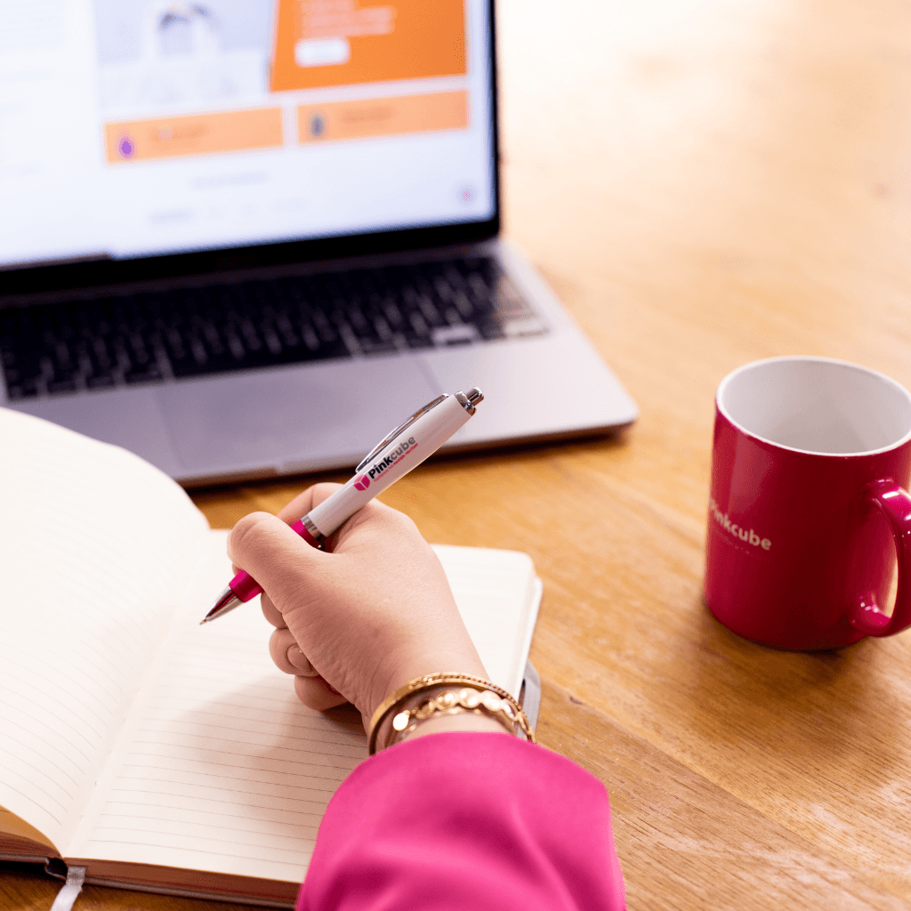 Hand die met een Pinkcube-balpen in een open notitieboek schrijft, met op de achtergrond een laptop en een roze Pinkcube-mok op een houten tafel.