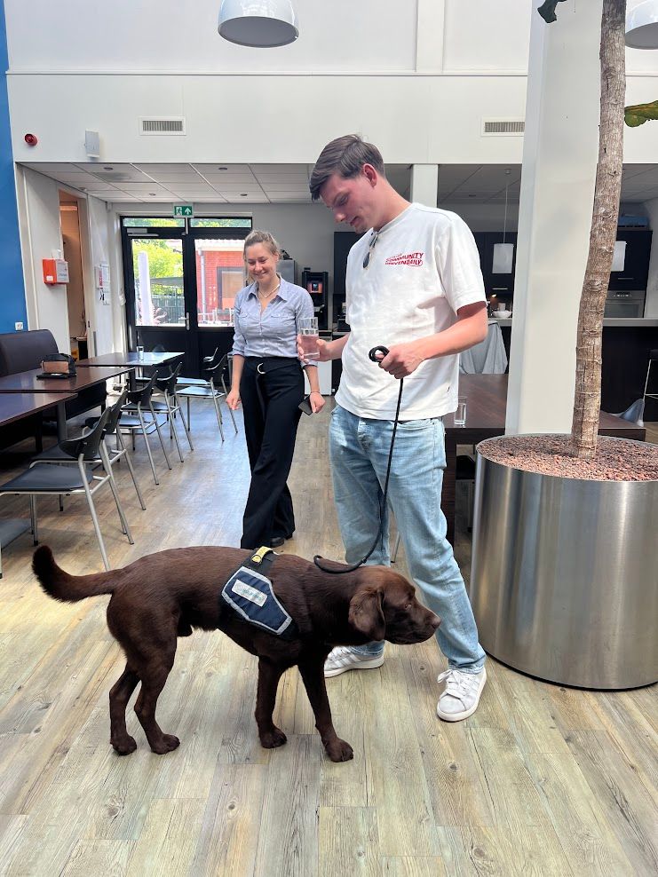 Collega in een bedrijfsruimte houdt Hulphond Kees aan de lijn terwijl hij een glas water vasthoudt. Op de achtergrond loopt een vrouw glimlachend mee in de open, licht ingerichte kantine.